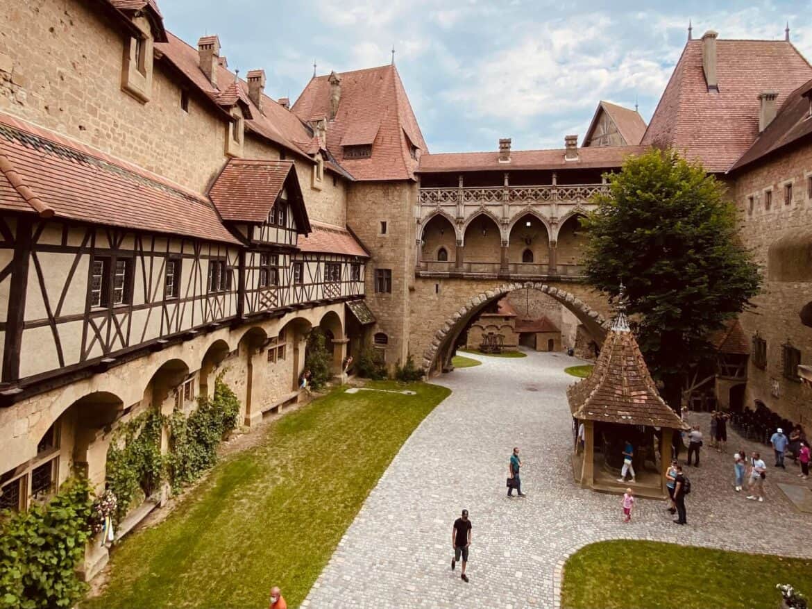 Blick in den Hof von Burg Kreuzenstein. Ein Weg in der Mitte. Links viele Fenster und ein Vordach. Unten im Hof ein überdachter Brunnen. Gerade aus sieht man ein Tor.