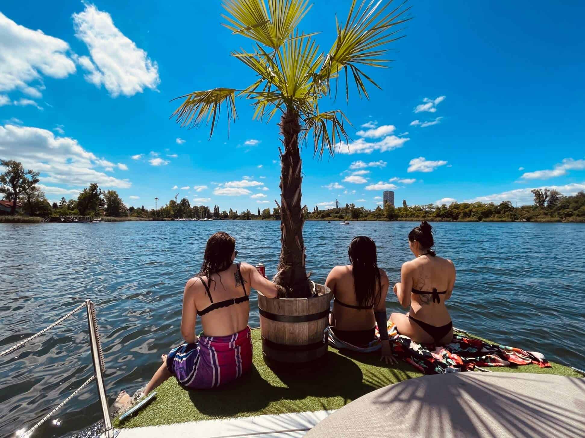 Drei Mädels sitzen am Rande eines Bootes. Am Boot eine Palme im Topf. Aussicht auf die alte Donau. Wasser im Bild. Sehr sonnig.