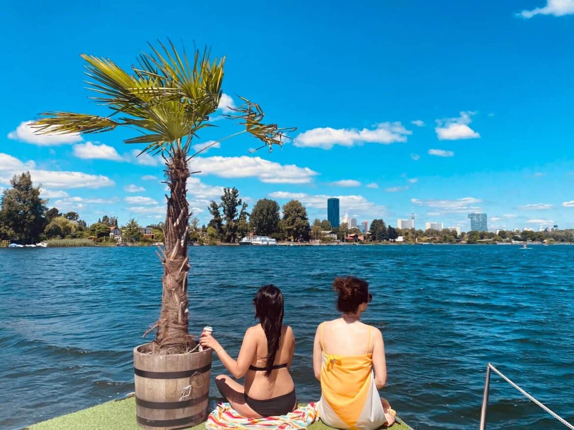 Zwei Mädels sitzen am vorderen Rand vom Inseloot. Am Boot eine Palme im Topf. Aussicht auf die alte Donau. Wasser im Bild.