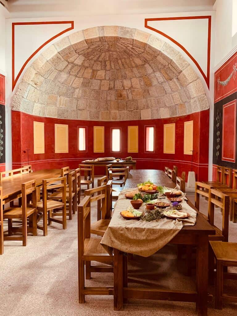 Tische und Stühle in einer ehemaligen (nachgebauten) Mensa der Römerstadt Carnuntum. Die Wände sind rot und es befindet sich am hinteren Ende eine runde Kuppel. Der Tisch ist mir einem Tischtuch dekoriert, ebenso wie mit Tellern und künstlich (fake) Speisen als Deko.