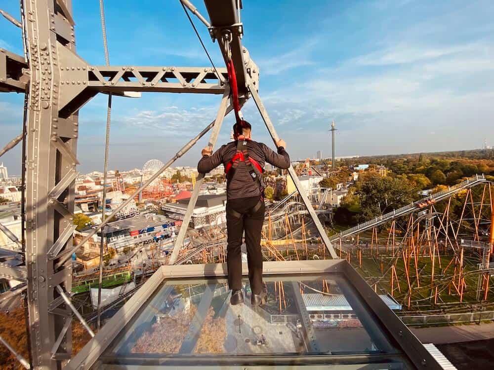 Mann von hinten gesehen steht auf der gläsernen Plattform 9 des Wiener Riesenrads. Links ein Teil der Konstruktion. Ausblick über den Wiener Prater.