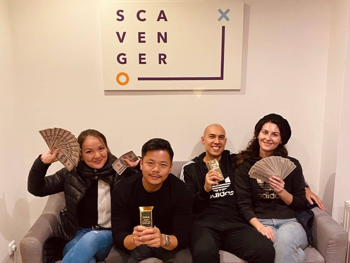 4 Personen sitzen auf einer Bank bei Scavenger Escape und haben erfolgreich eine Schnitzeljagd gelöst. In der Hand haben sie Spielgeld und Goldbarren. Alle grinsen.