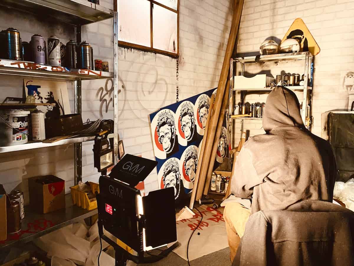 Mystery of Banksy Austellung in Wien. Rechts ein bedeckter myteriöser Mann mit Hoodie von hinten in einem Raum voller Banksy Kunstwerke.