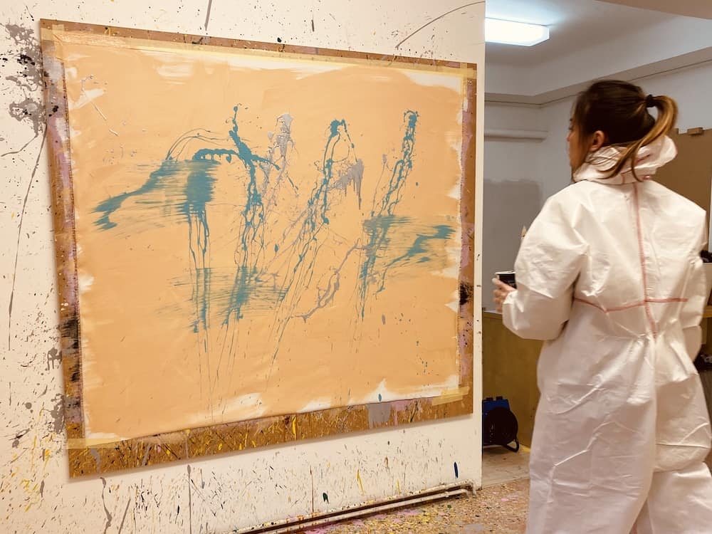 Leinwand mit gelben Hintergrund und blauen Farbspritzern. Frau mit weißen Schutzanzug übt Action Painting aus.