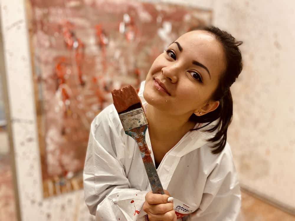 Frau mit Pinsel in der Hand sieht glücklich und zufrieden vom Action Painting aus. Im Hintergrund ihr gemaltes Bild verschwommen zu sehen.