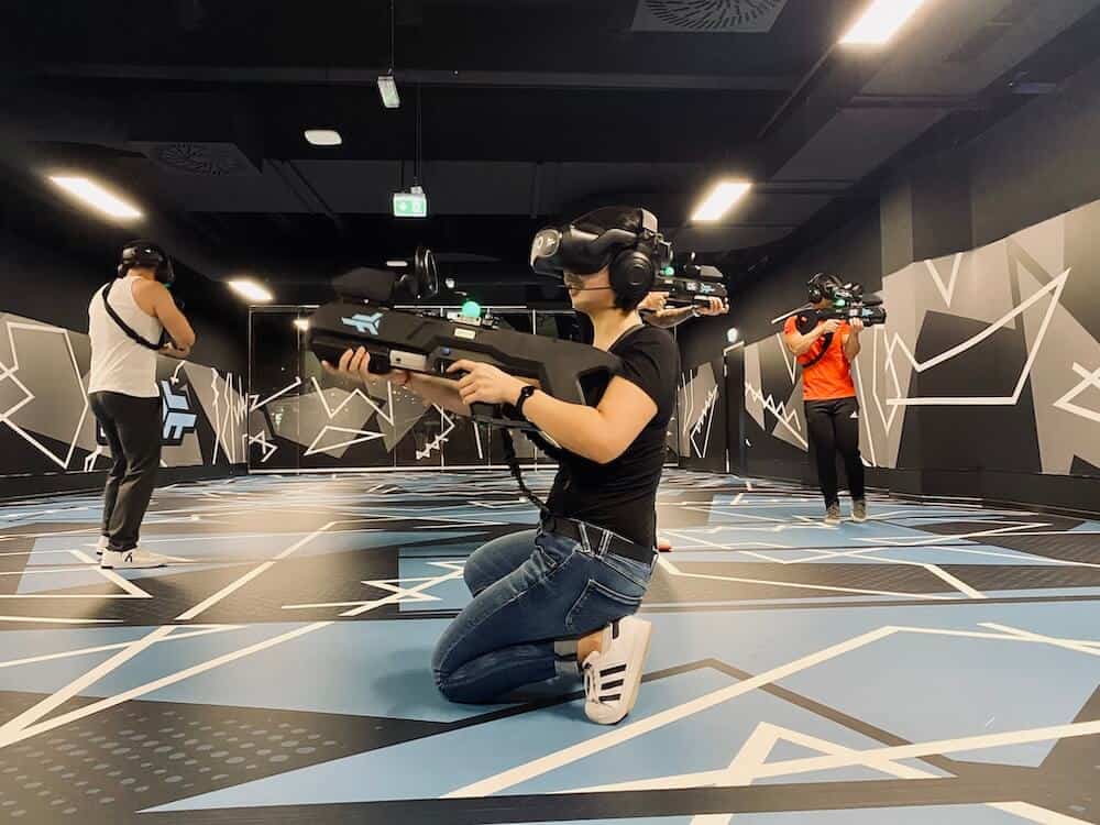 Mädchen kniend mit VR Brille und Spielgewehr für ein Virtual Reality Gaming. Andere Mitspieler im Hintergrund