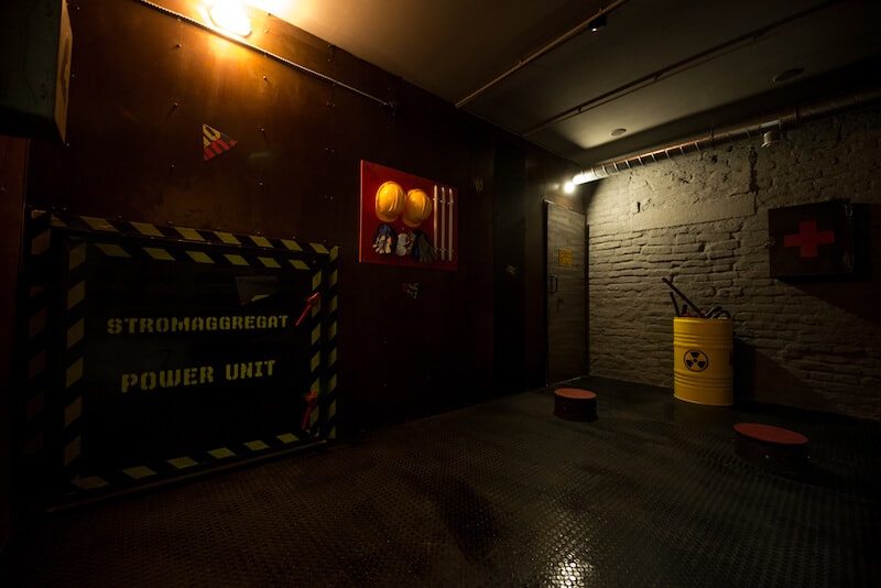 Neuer Escape Room inm Wien. Dunkler Raum. Mit gelben Gefahren Fass auf der rechten Seite und Schutzhelme an der Wand. Der Raum soll ein Militätlabor nachstellen.