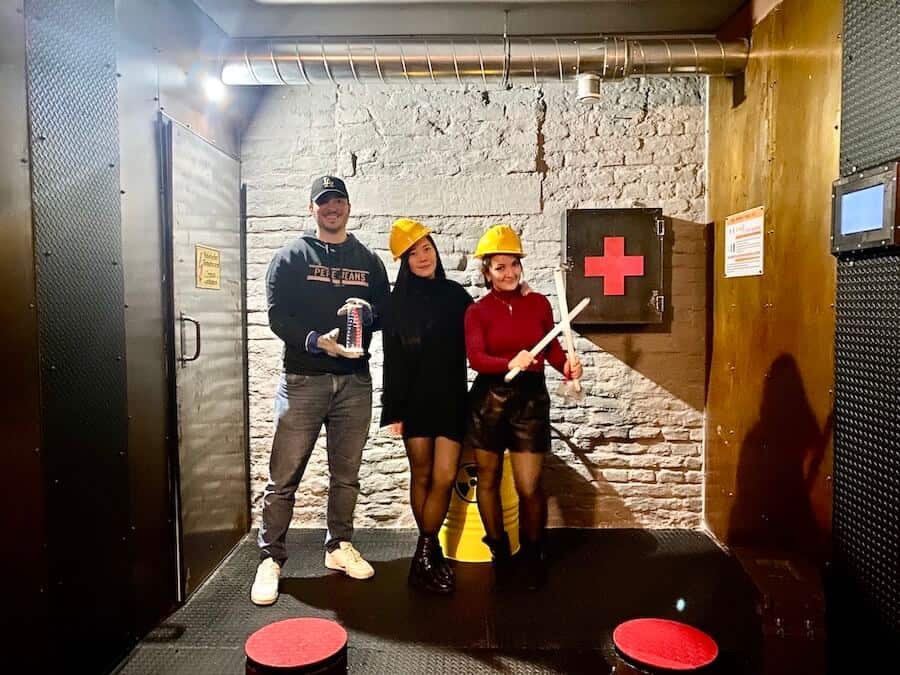 3 Spieler:Innen spielen einen neuen Escape Room. Posieren mit gelben Helmen und Leuchtstoffröhren. Der Raum ist dunkel und erinnert an ein Militärbunker