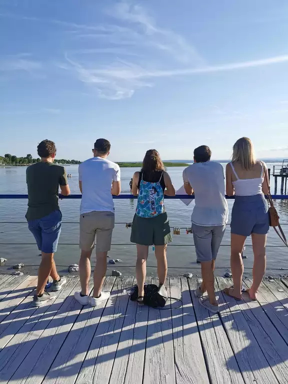 5 Leute von hinten. Stehen an einem Steg vorm See.