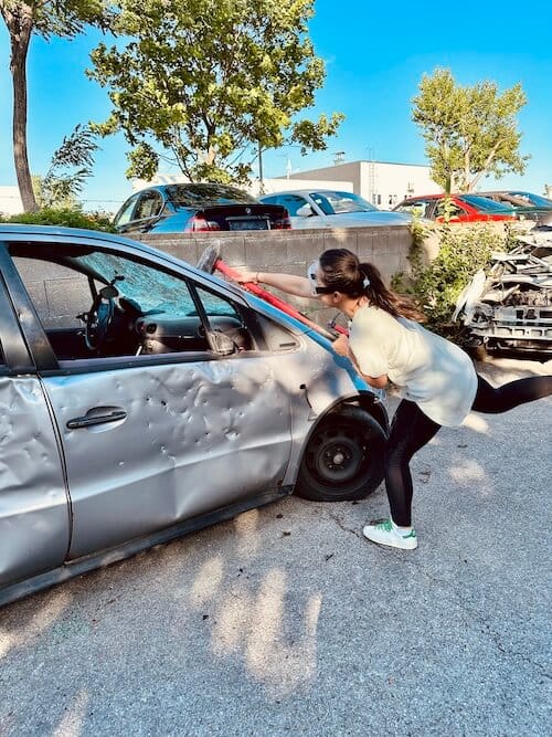 Silbernes Auto und Frau, die mit einem Vorschlaghammer auf die Windschutzscheibe schlägt.