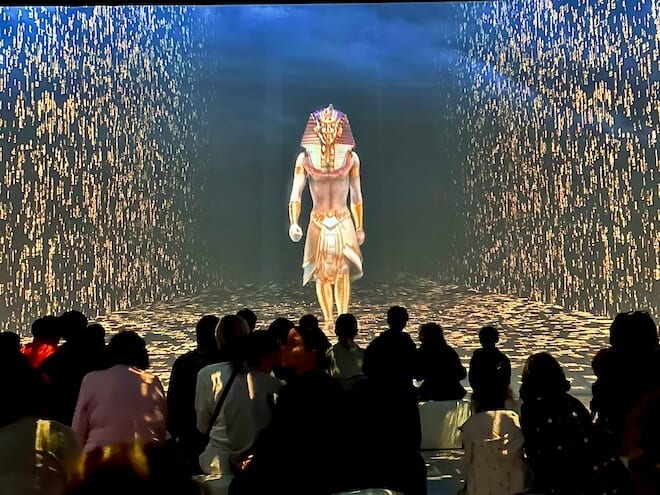 Multidimensionaler Raum der Tutanchamun Immersive Ausstellung. Auf er 360 Grad Leinwand ist ein Pharao zu sehen. Menschen sitzen auf Blöcken in der Mitte und beobachten das Szenario.