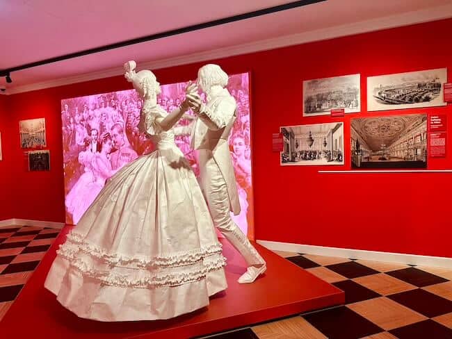 Weiße Skulpturen, die den Walzer tanzen. EIne Frau im Kleid und Mann im Frack in einem komplett roten Raum.