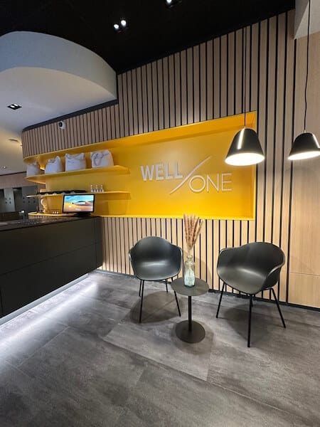 Eingansbereich des Well-One. Zwei graue Stühle und einen Tisch vor gelb-holzigem Hintergrund und einen Teil der Rezeption.