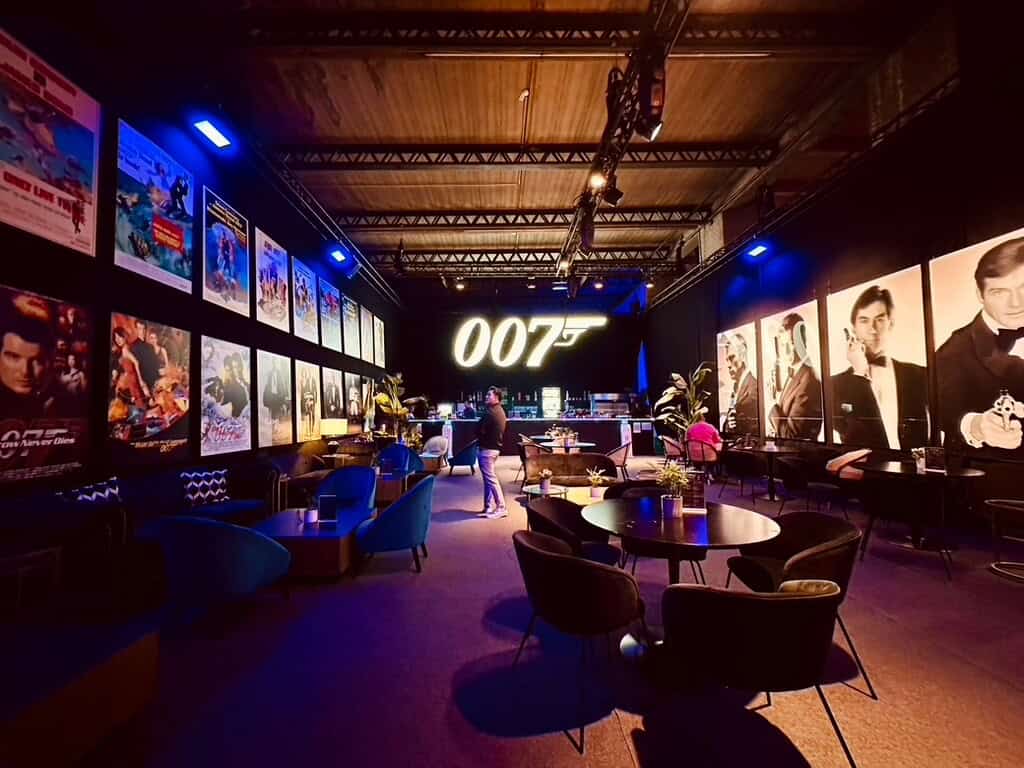Lounge der 007 James Bond Ausstellung. Links Filmplakate der FIlme an der Wand. Rechts alle Bond Schauspieler. In der Mitte Lounge Möbel und hinten eine Bar mit der leuchtenden Aufschrift 007.